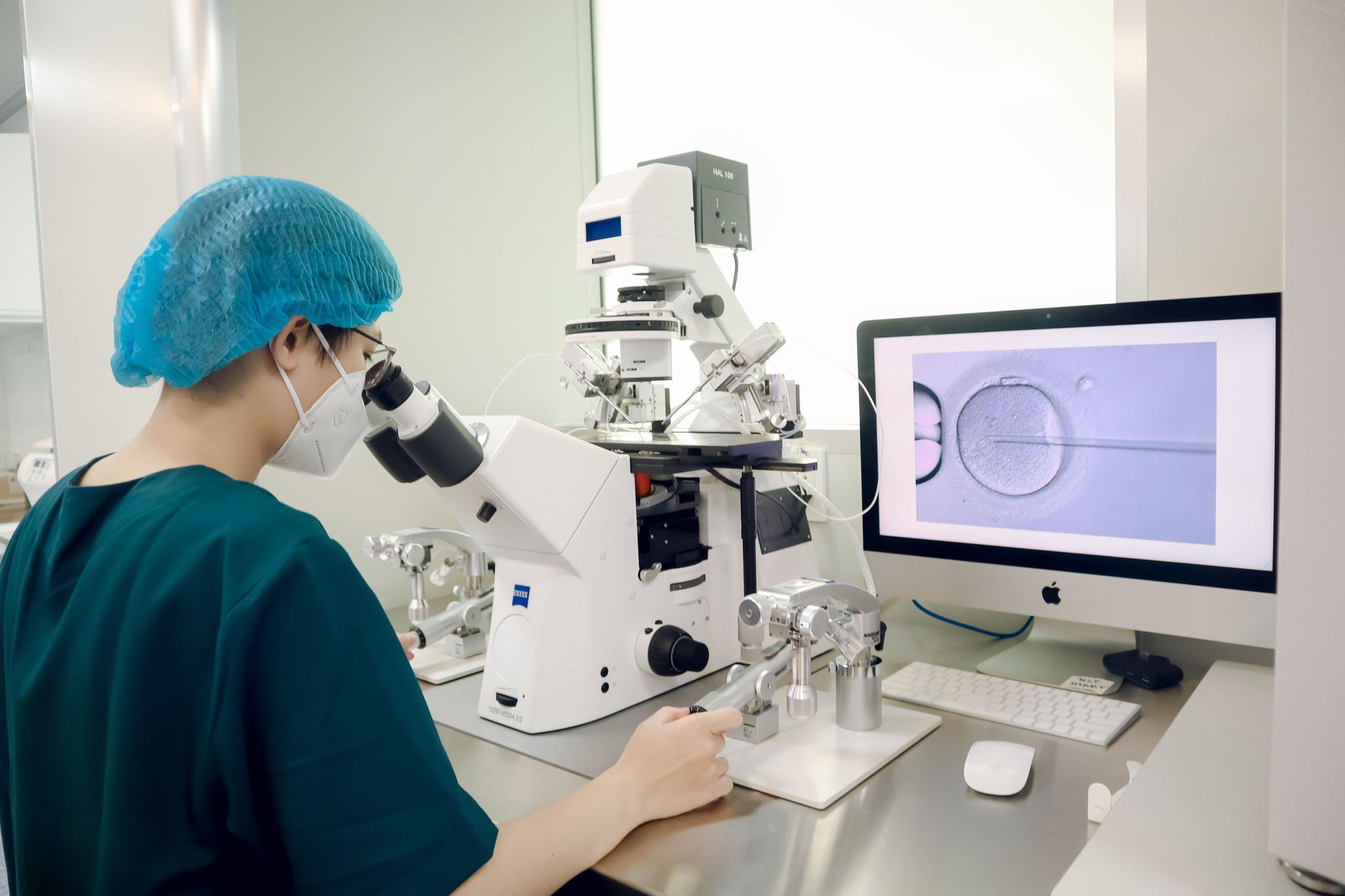 Un technicien de laboratoire portant un masque et une charlotte, observant un microscope dans un laboratoire, avec un écran affichant une image agrandie d'une cellule.